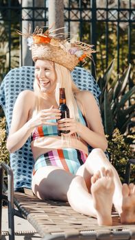 Woman enjoying a sunny day by the pool in a vibrant swimsuit and straw hat, holding a beer.