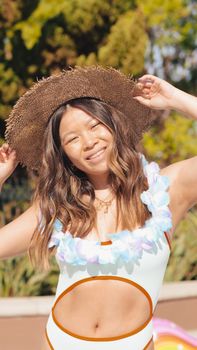 Smiling woman in swimsuit with sun hat and floral garland enjoying a sunny poolside moment.