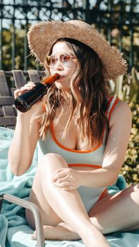Woman in a swimsuit enjoying a drink on a sun lounger by the pool, embracing summer vibes.
