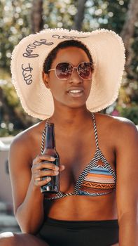 Black woman in bikini enjoying summer sun, holding a bottle and wearing sunglasses and sun hat.