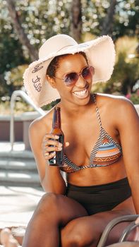 A joyful woman in a bikini relaxes with a beer at a sunny poolside, wearing a sun hat and sunglasses.