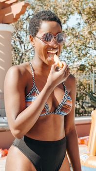 A joyful woman in a striped bikini enjoys a light snack by the pool on a sunny summer day.