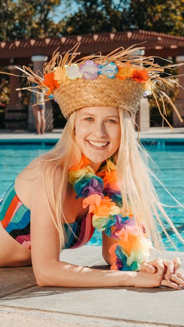 Cheerful young woman in vibrant lei and straw hat enjoying a sunny day by the pool.