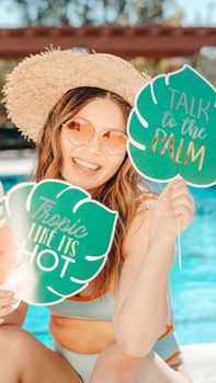 Woman enjoying a sunny poolside party, wearing a sun hat and holding playful signages.