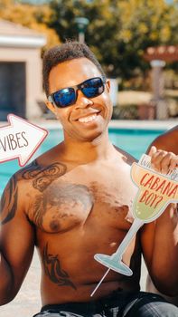 Man with tattoos holding fun signs at a summer pool party. Sunglasses and joyful expression.