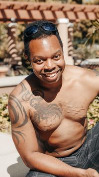 Smiling tattooed man relaxing poolside in sunny summer weather, wearing sunglasses.