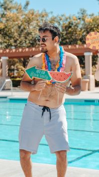 Happy man at a summer pool party holding festive props and wearing sunglasses and a colorful garland.