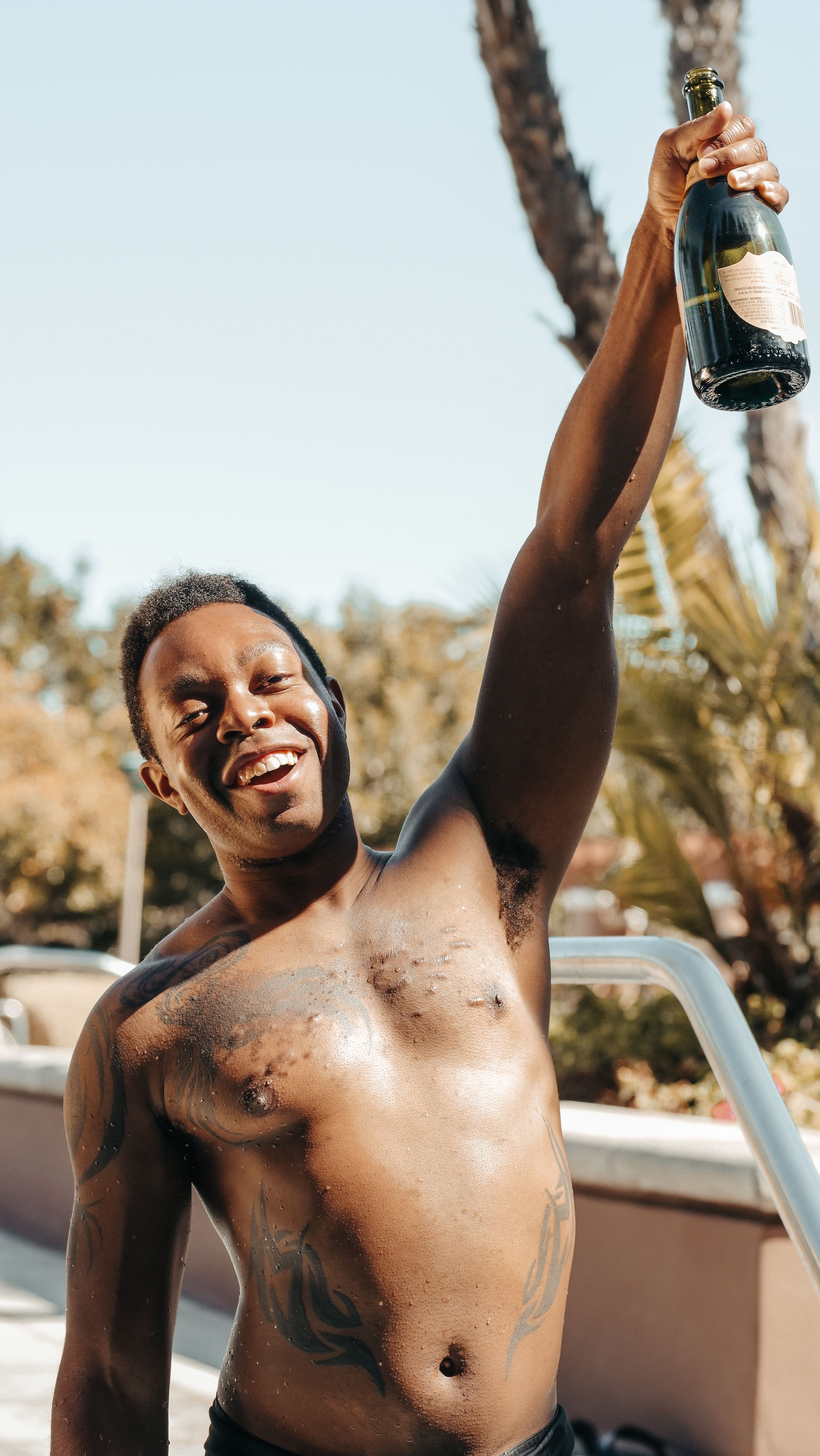 Smiling shirtless man holding a champagne bottle at a vibrant summer pool party.