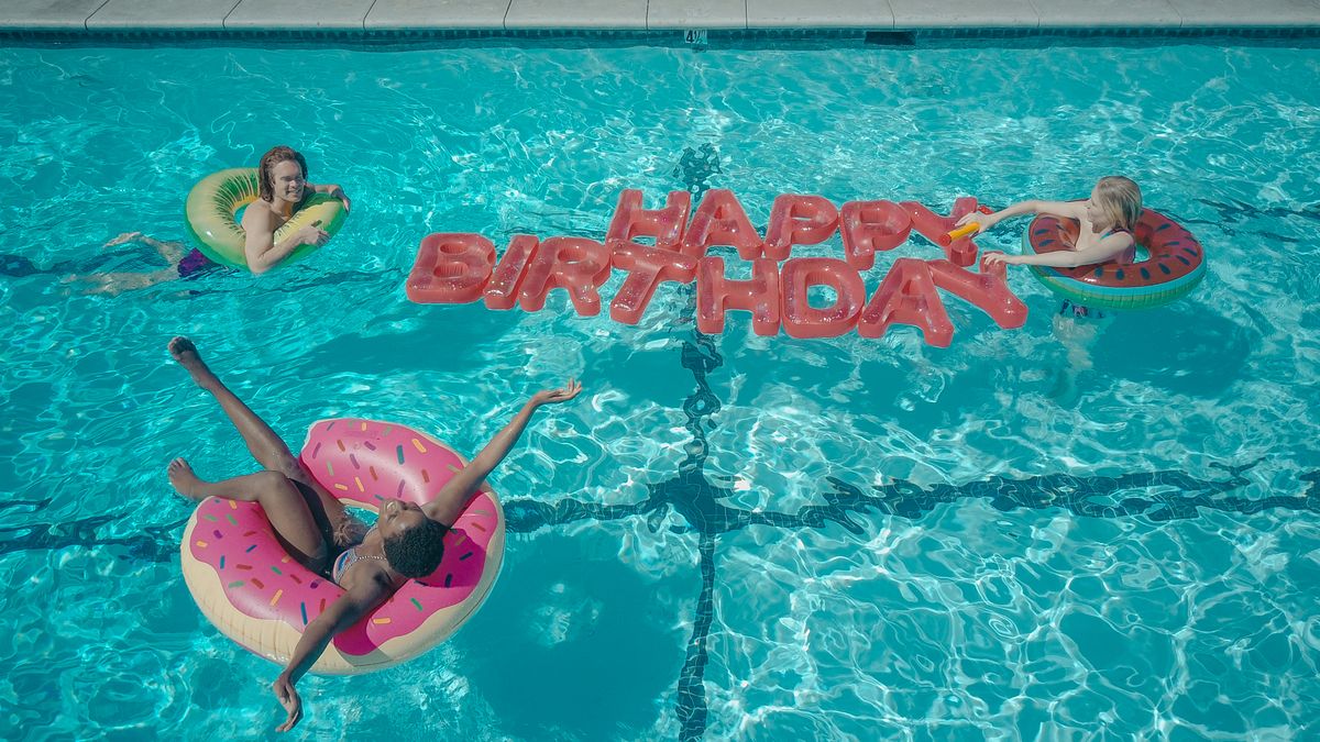 Three friends enjoying a birthday celebration at a pool with colorful inflatables and a happy birthday message. Stock Photo.