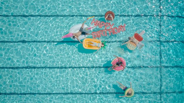 Top view of friends enjoying a summer pool party with floats and 'Happy Birthday' balloons.