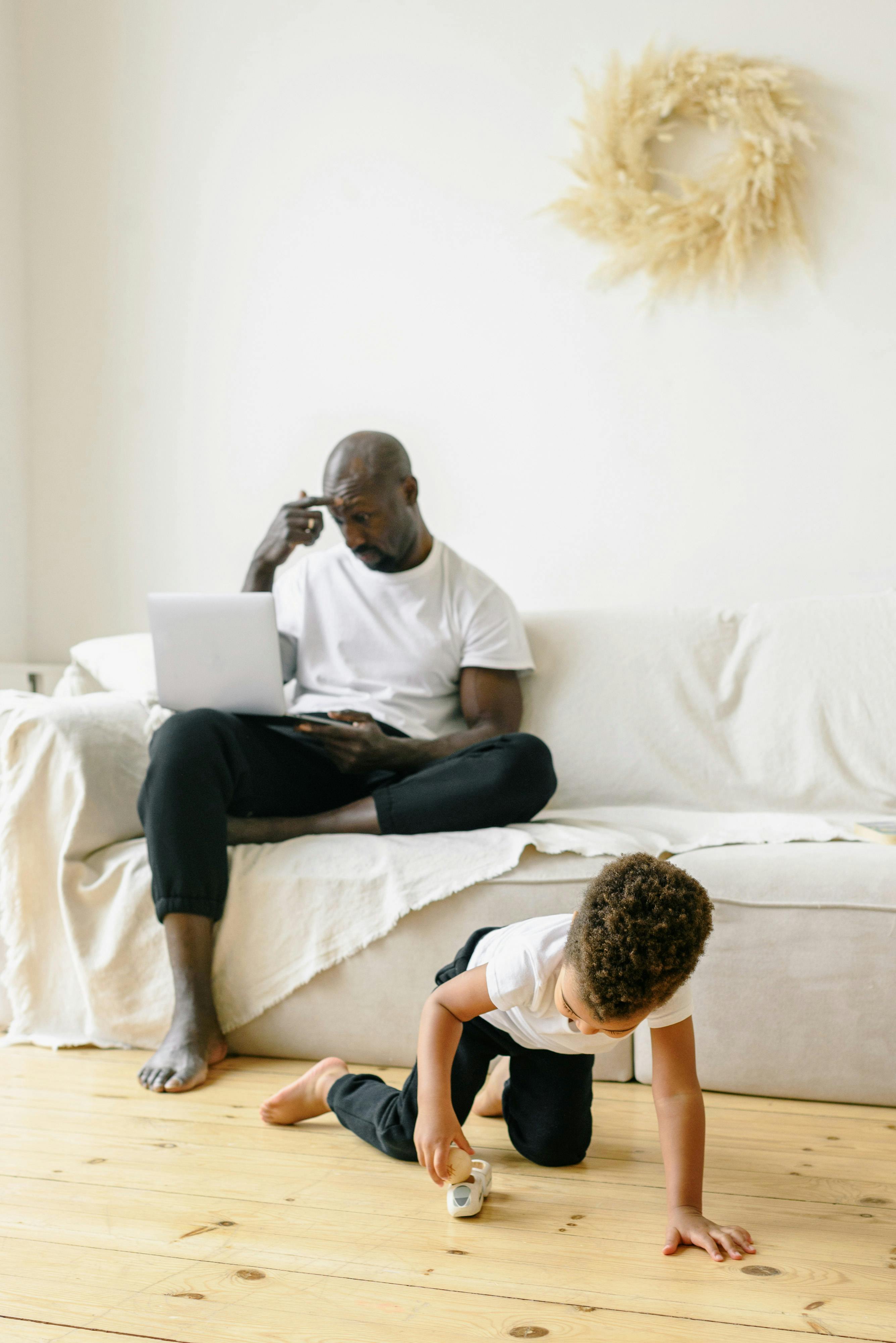 little boy playing with a toy and his father sitting on a couch using laptop