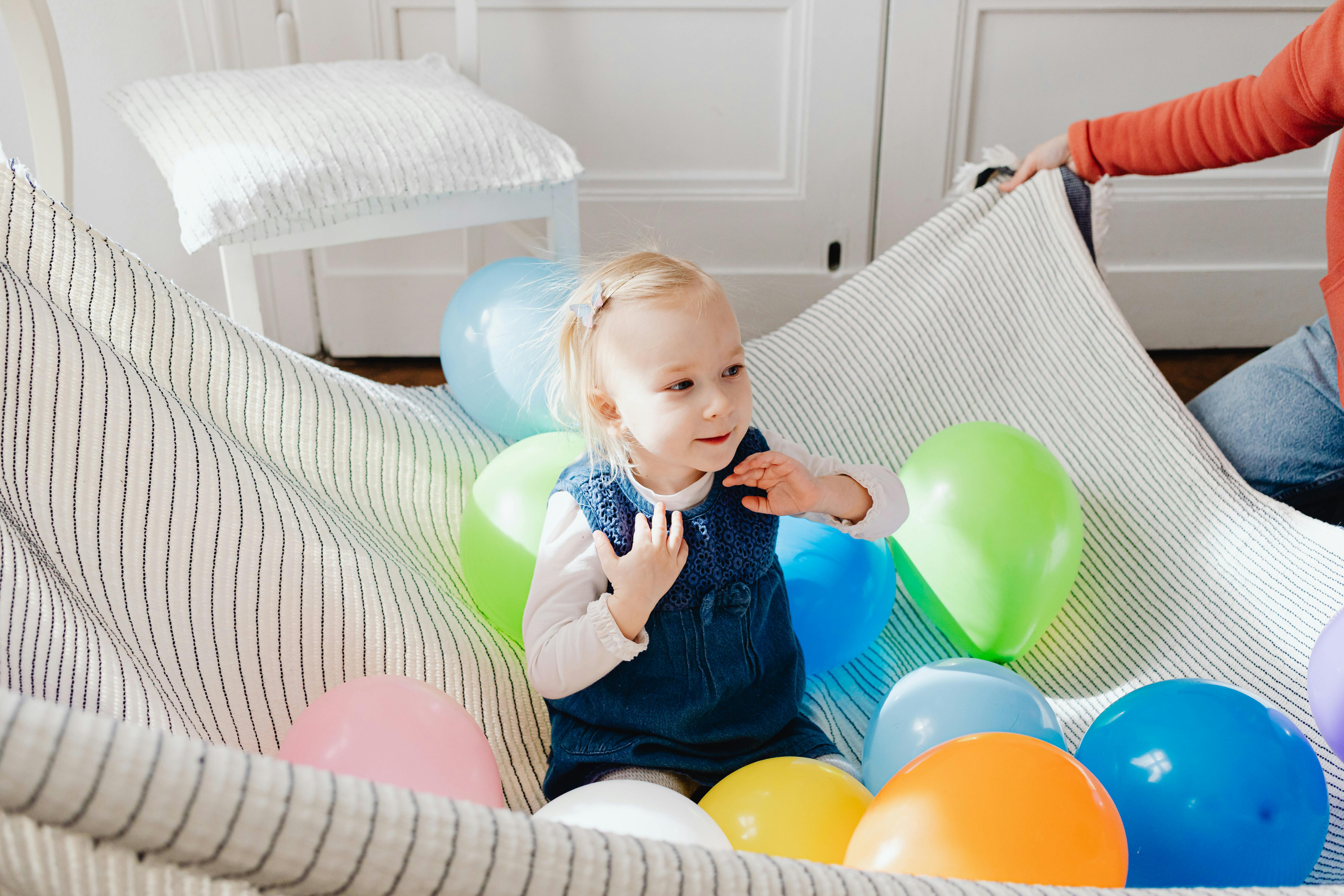 A young girl in a striped hammock surrounded by colorful balloons, enjoying playful indoor fun.