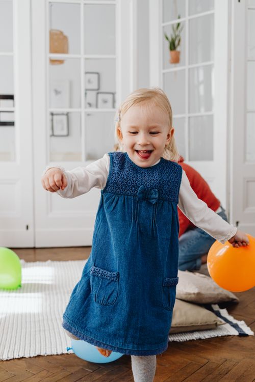 Free Happy toddler in a blue dress playing with colorful balloons indoors. Stock Photo
