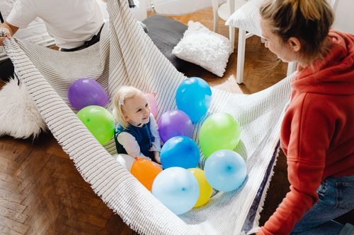 Free A young child happily playing with colorful balloons on a blanket indoors, surrounded by adults enjoying the moment. Stock Photo