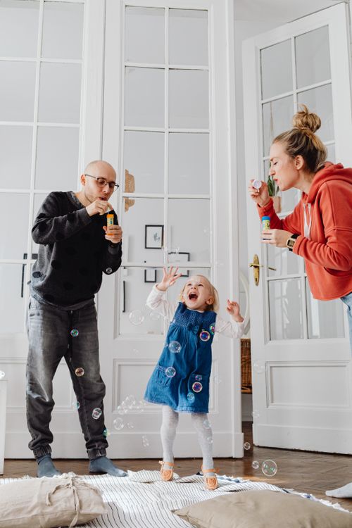 Free Parents joyfully blowing bubbles with their child, creating a warm and happy family moment indoors. Stock Photo