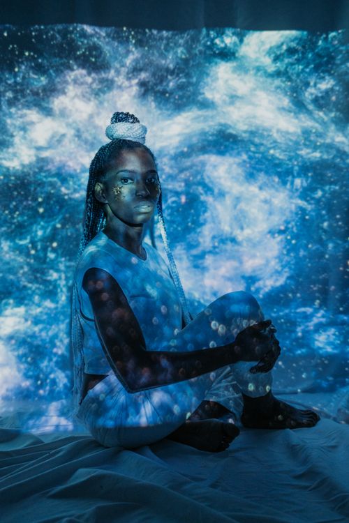 Free Woman with braided hair sitting against a cosmic galaxy backdrop with blue starry projections. Stock Photo