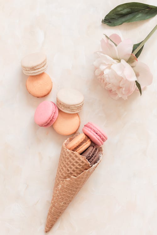 Free Overhead sweet macaroons in waffle cone and scattered on white table near fragrant delicate flower Stock Photo