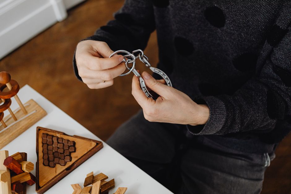 Top view of hands solving metal puzzle, surrounded by wooden brain teasers on a table.