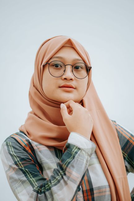 Portrait of a young woman wearing a headscarf and glasses in a studio setting with a white background.