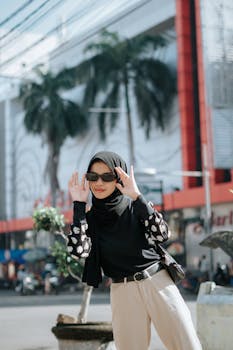 Confident woman in hijab and sunglasses smiling and posing in a vibrant city street.