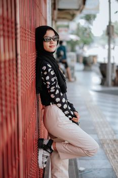 A fashionable young woman wearing sunglasses and a headscarf, stylishly leaning against a red wall in urban setting.