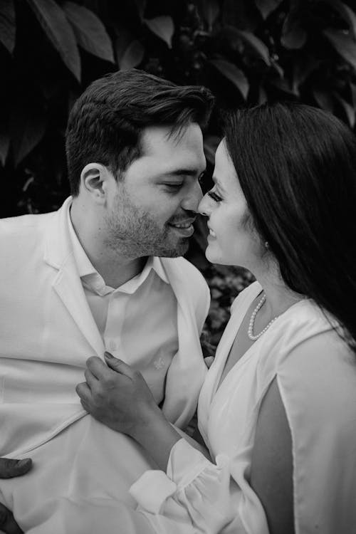 Black and white of ethnic smiling couple in white clothes looking at each other tenderly while standing in park