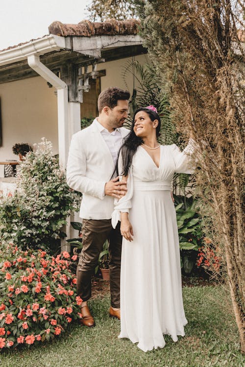 Free Happy ethnic newlywed couple in elegant wedding clothes looking at each other while standing near tree in garden with blossoming bushes Stock Photo