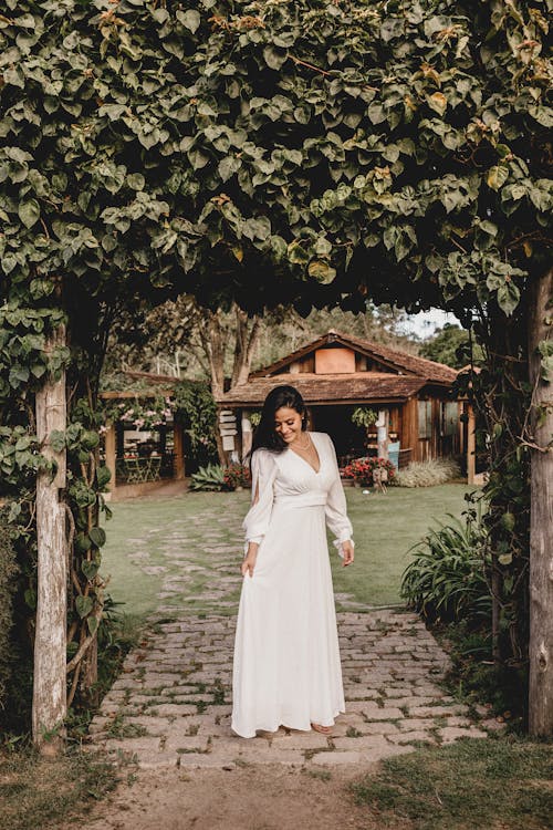 Smiling Indian woman in white dress with long hair standing near green plants against background of cottage