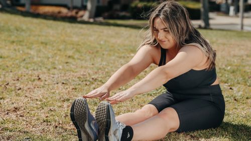 Free Caucasian woman stretching on grass in athletic wear, enjoying a sunny outdoor workout. Stock Photo