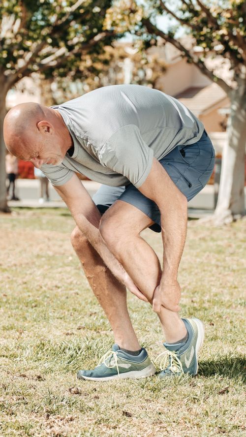 Free Man holding his injured ankle outdoors in a park, highlighting running-related pain or sport injury. Stock Photo