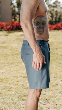 Shirtless man with tattoo stands outdoors in shorts, showcasing a fit lifestyle.