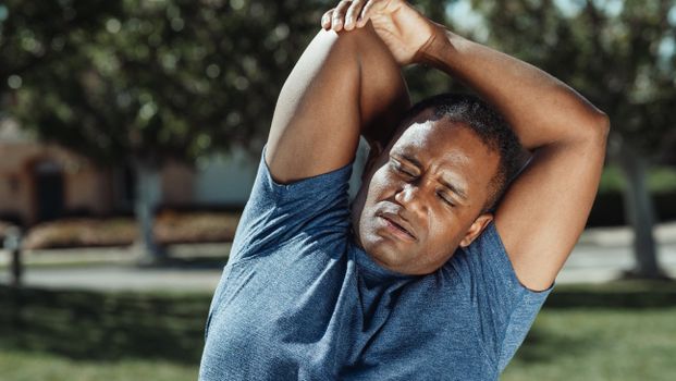 A man in a blue shirt performing a stretching exercise outdoors, promoting healthy living and fitness.