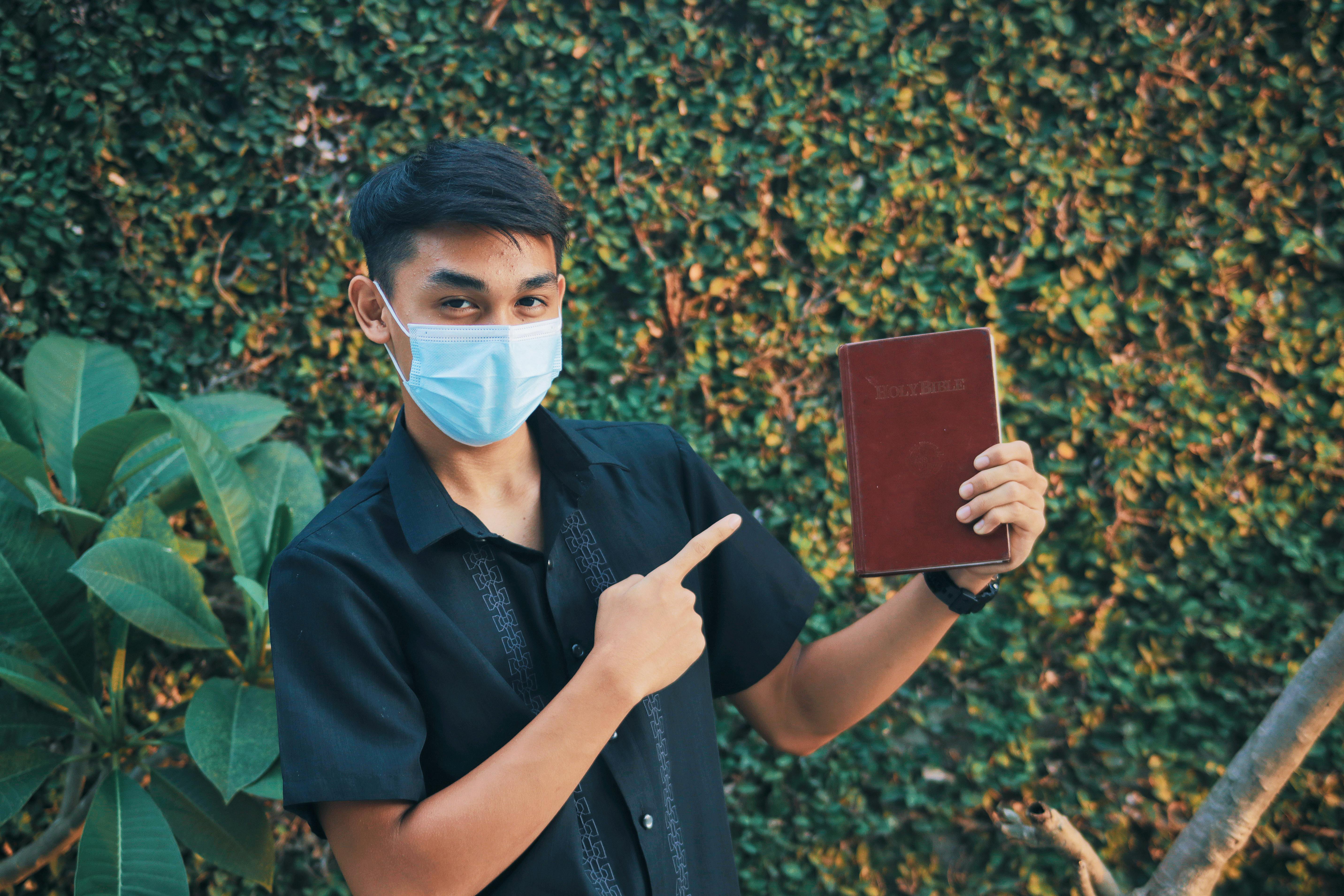 A young man wearing a face mask holds a Bible, pointing at it with a blurred green background.