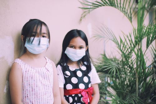 Free Two young girls with face masks posing by a wall with plants outdoors. Stock Photo