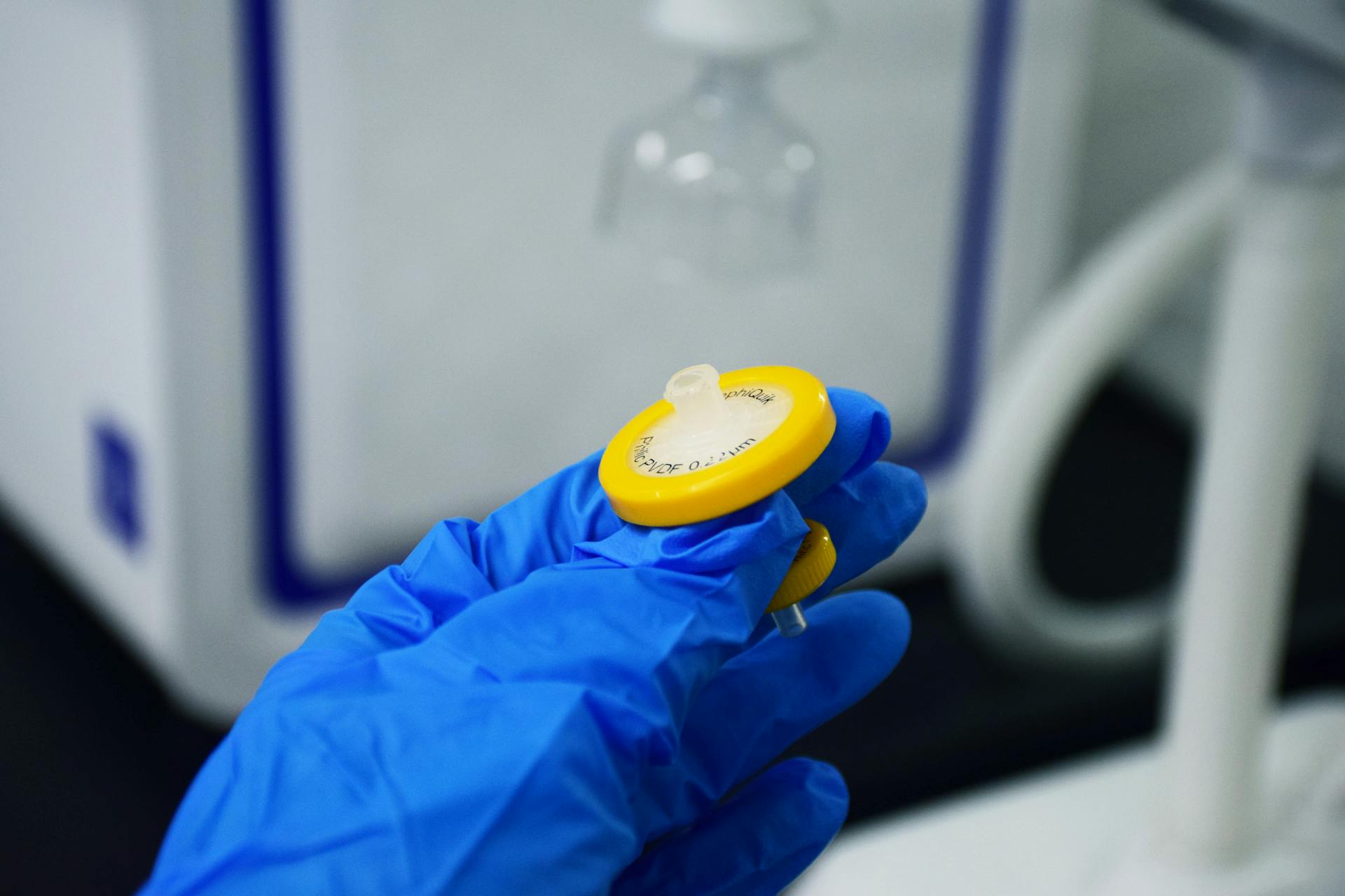 Detailed close-up of a hand in a blue glove holding a laboratory syringe filter indoors.