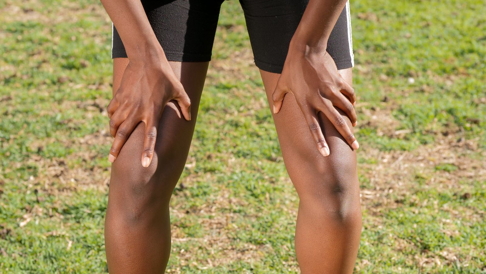 A Black person in athletic shorts grips both knees with both hands while standing on grass, a common posture when knee pain flares after activity.