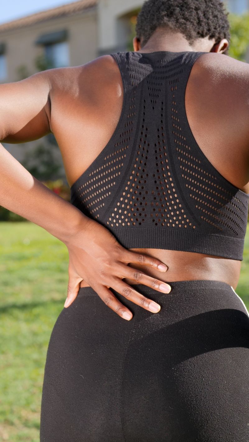 A Black woman in athletic wear presses one hand to her lower back, the lumbar spot where sciatic nerve pain and low back pain both begin.