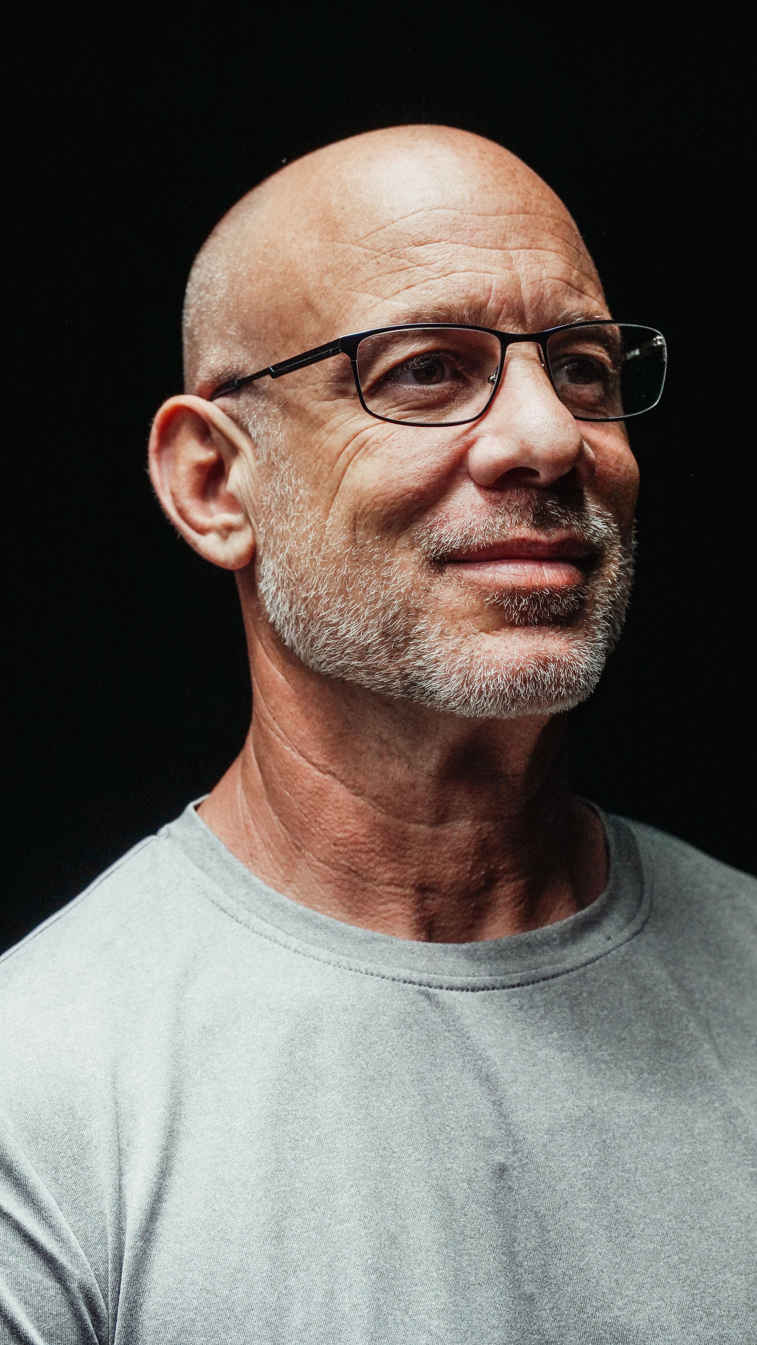 Portrait of a bald man with glasses smiling warmly in a gray shirt against a dark background.