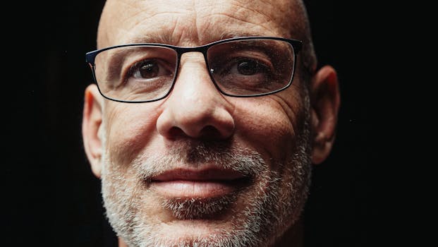 Close-up portrait of a smiling bald elderly man with glasses against a dark background.