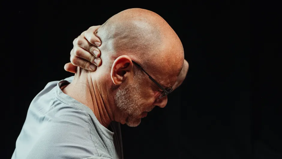 Homme senior de profil, tête penchée en avant, main serrant la nuque, geste typique de la cervicalgie qui accompagne un acouphène cervical