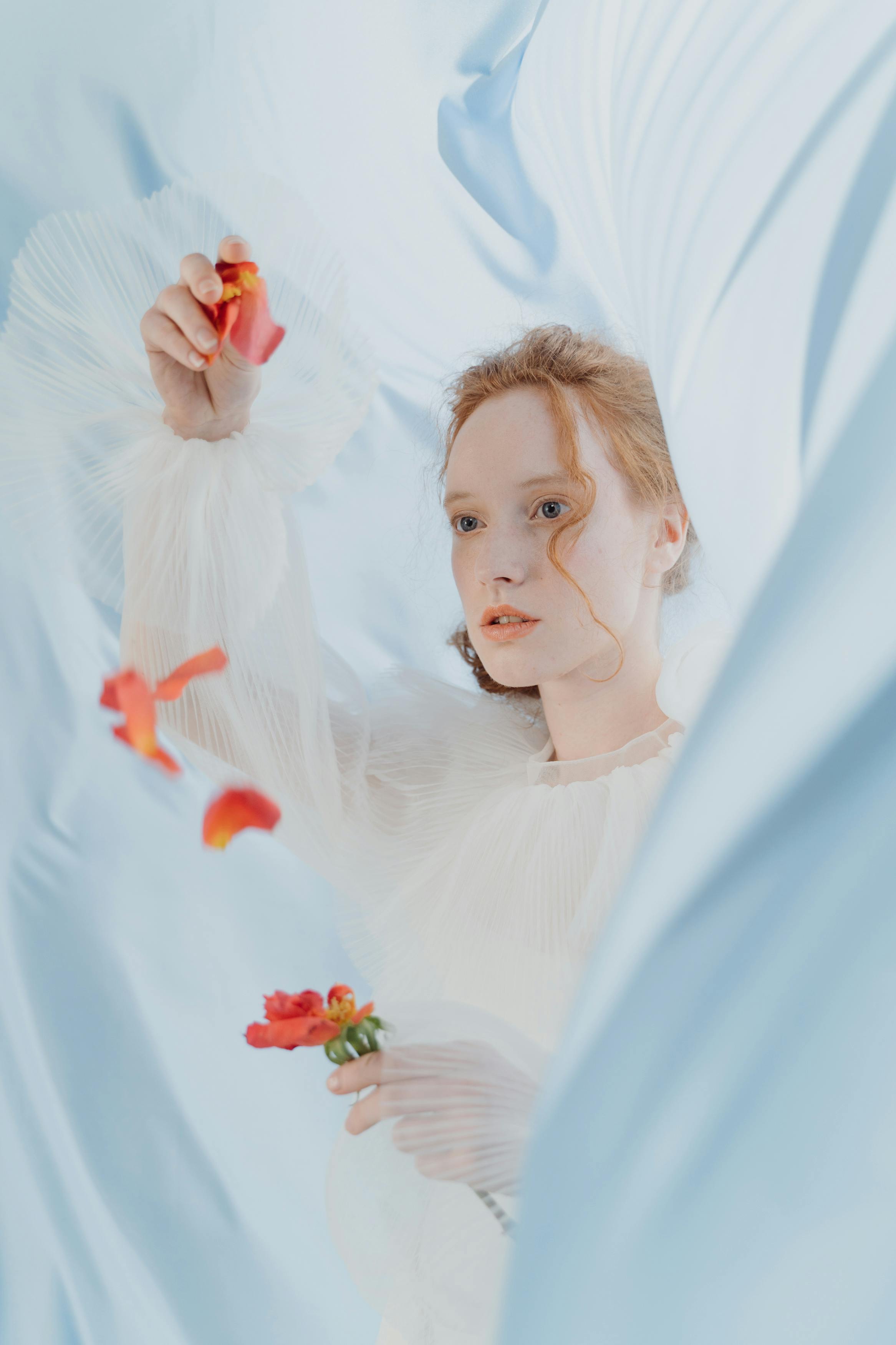 Elegant woman with red hair holding flower, surrounded by flowing fabric and petals.