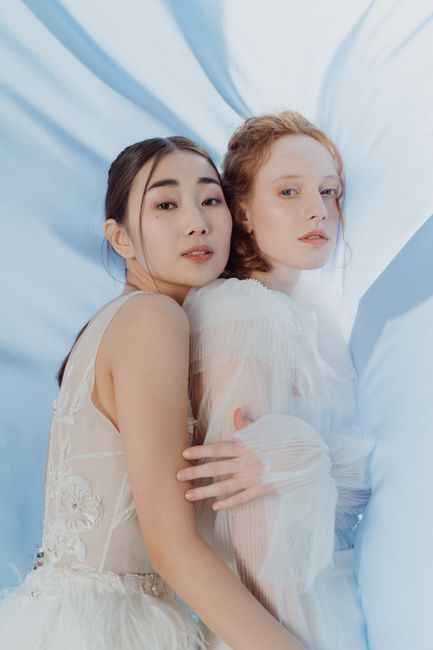 Two women in elegant wedding gowns gently embracing against a soft blue backdrop.