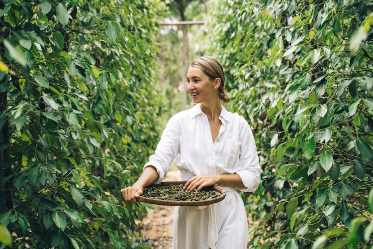 Woman In White Long Sleeves Dress Looking At The Green Plants While Holding A Round Winnower