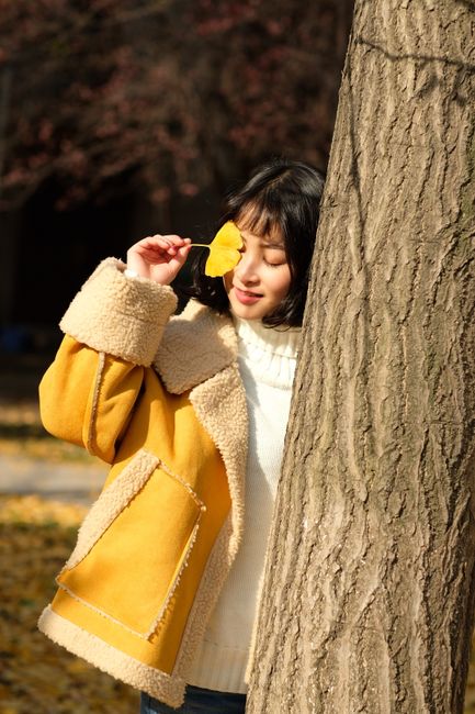 A woman in a yellow coat enjoys a sunny fall day with a leaf in hand.