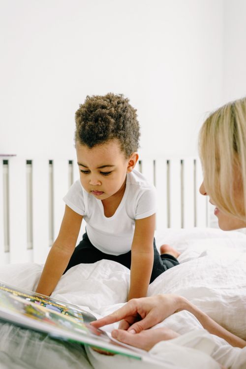 Free A mother and her young son sharing a bonding moment while reading a book together in a bright bedroom. Stock Photo
