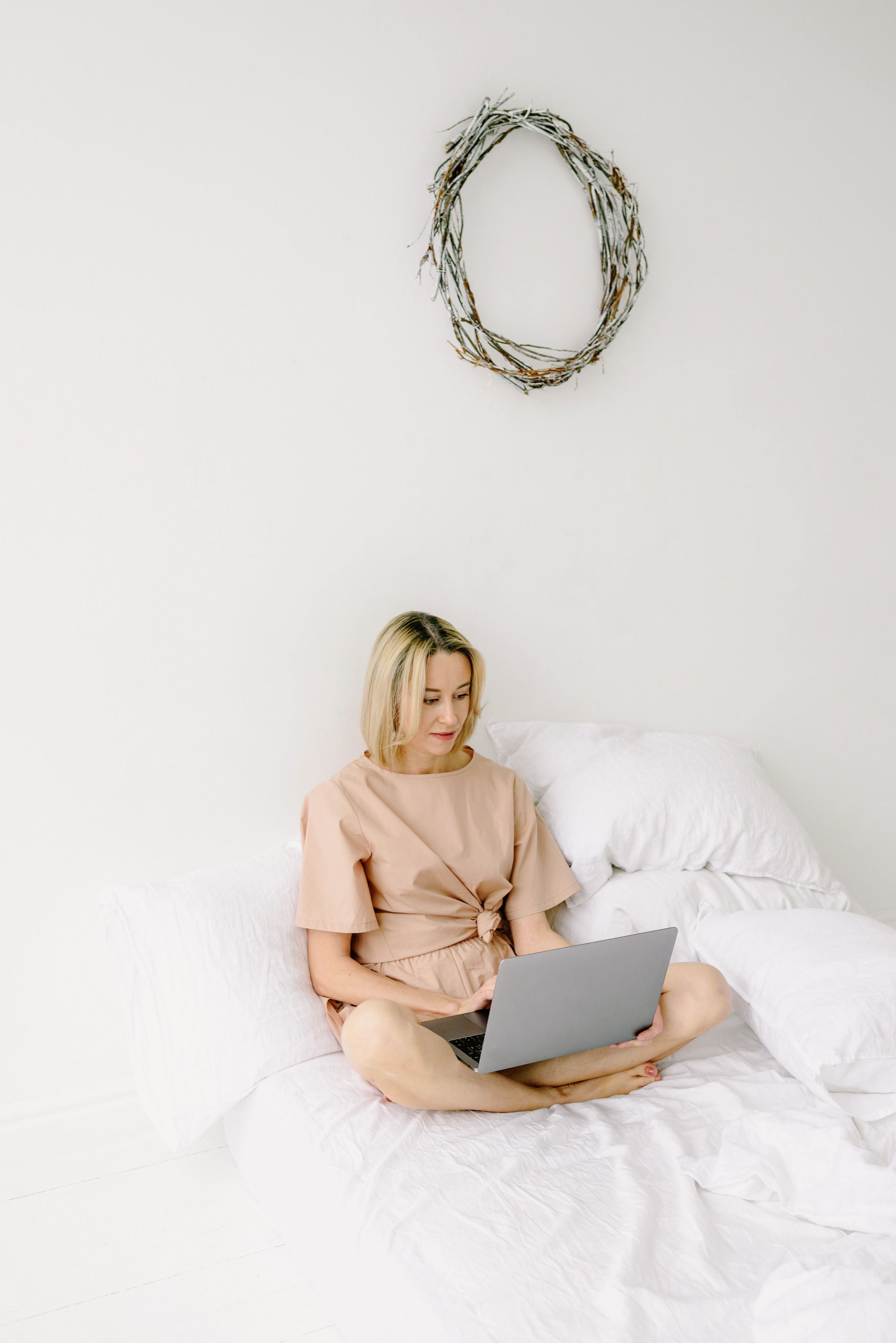 Woman working from home in a bright, minimalist bedroom setting with a laptop.