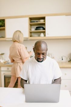 Adult man working on a laptop in a contemporary kitchen while a woman prepares food in the background.