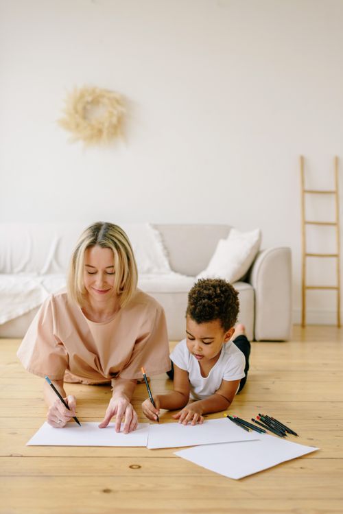 Free A mother and child drawing with pencils on paper while lying on the wooden floor in a cozy living room. Stock Photo