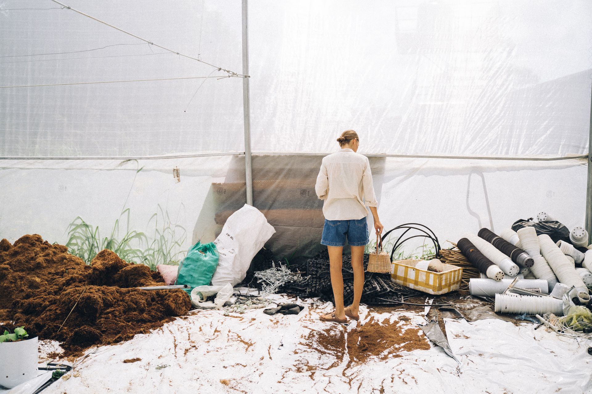 A woman stands in a greenhouse setting, organizing gardening tools and materials on a sunny day.
