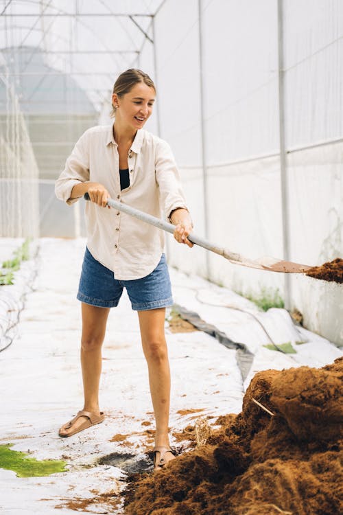 A Woman in White Long Sleeves and Denim Shorts Shoveling Soil